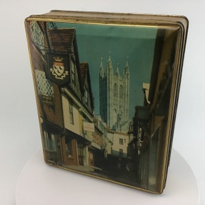 May include: A rectangular tin box with a gold trim and a scenic image of a European town. The image features a tall cathedral and buildings along a street. The box is slightly angled, showing its side.