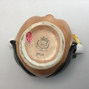 May include: A brown ceramic pitcher with a black mustache and a yellow flower. The pitcher has a white base with a gold design and the text "ESD Taiwan Hand Painted 21728".