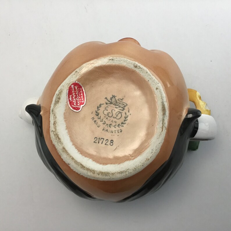May include: A brown ceramic pitcher with a black mustache and a yellow flower. The pitcher has a white base with a gold design and the text "ESD Taiwan Hand Painted 21728".