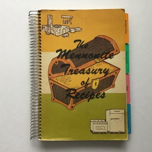 May include: A yellow, green, and orange cookbook with a spiral binding. The cover features an illustration of an open treasure chest with the text "The Mennonite Treasury of Recipes".