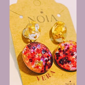 May include: A pair of two-part earrings featuring a small oval stud with gold leaf flakes and a larger circular drop pendant filled with red, pink, and iridescent glitter. The earrings are attached to a tan cardboard display card labeled NOIA VERA.