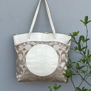 May include: A beige and white tote bag featuring a repeating monogram pattern on the fabric body. The bag has a large white circular leather patch in the centre, white leather trim at the top, and two long white leather shoulder straps.