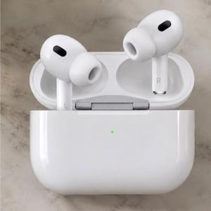 May include: A pair of white wireless earbuds with black oval accents on the outer surface, each marked with L or R, resting inside an open white rectangular charging case that features a small green status light on the front exterior surface.
