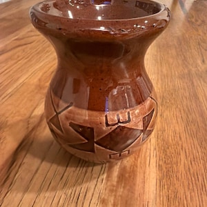 Può includere: Un vaso in ceramica marrone fatto a mano con un bordo ampio e svasato e un corpo bulboso. Il vaso presenta un design geometrico in una tonalità più chiara di marrone al centro. Il vaso viene probabilmente utilizzato per scopi decorativi.