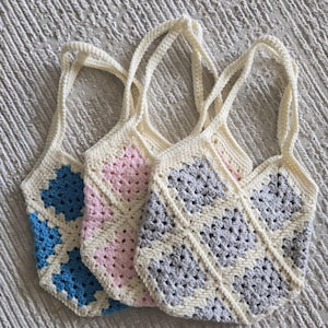 May include: Three handmade crochet tote bags in blue, pink, and gray. Each bag features a cream-colored border and handles, with a geometric pattern in the center. The bags are displayed on a textured surface.