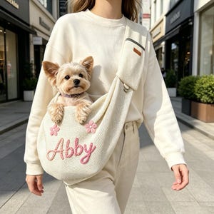 May include: A cream-coloured knit pet sling carrier featuring a wide shoulder strap and a small zipped pocket. The bag is decorated with two pink embroidered flowers and the name Abby stitched in pink yarn, containing a small tan dog.