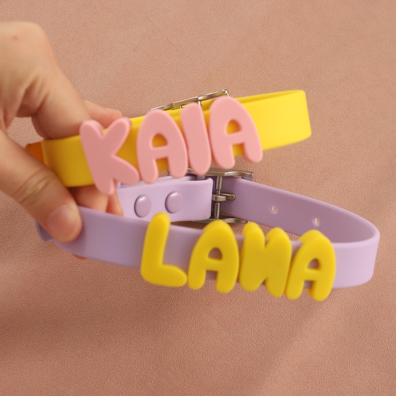 May include: Two silicone collars with metal buckles. The yellow collar features the name KAIA in pink letters, while the lavender collar displays the name LANA in yellow letters. Both collars have adjustable holes and a smooth, matte finish.