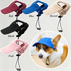 May include: A collection of pet visors in red, blue, apricot, black, and pink, each featuring an adjustable chin strap with a black toggle. A dog is shown wearing the blue visor, which has ear holes and a brim to shade the eyes from the sun.