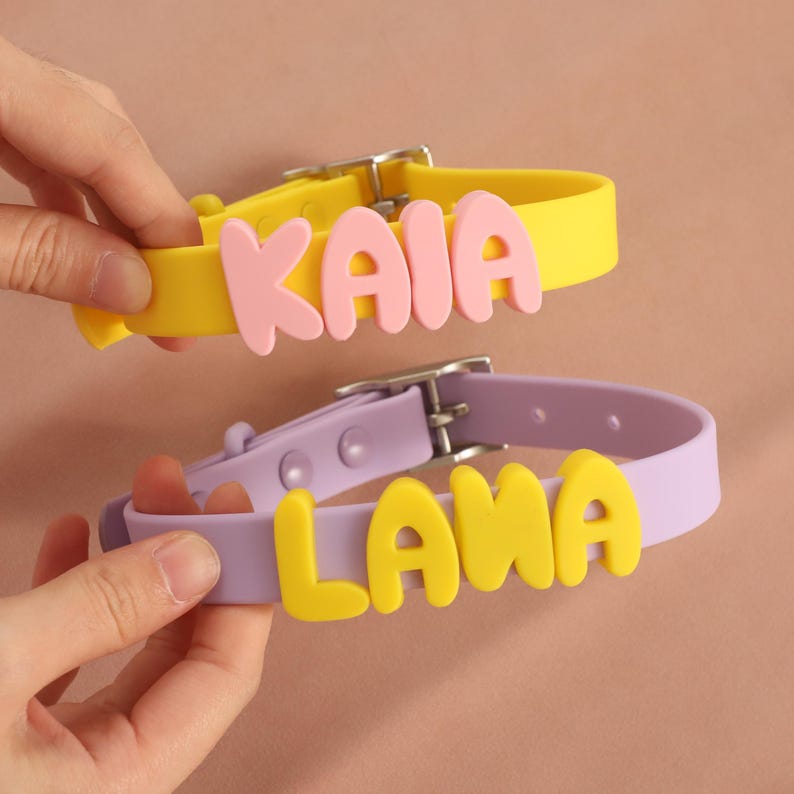 May include: Two silicone collars with metal buckles. The top collar is yellow with the name KAIA in pink letters. The bottom collar is light purple with the name LANA in yellow letters. Both collars feature adjustable holes and a smooth finish.