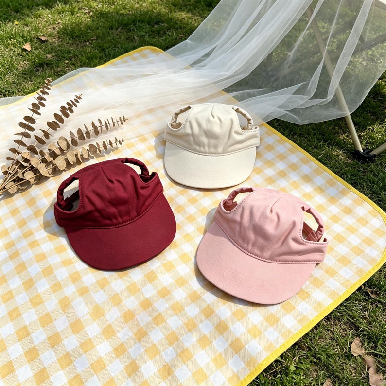 May include: Three pet sun visors in solid colors: one deep burgundy, one cream, and one soft pink. Each visor features a rounded brim and two side openings designed to accommodate ears, providing a secure fit for a pet's head during outdoor activities.
