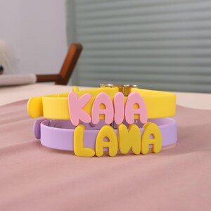 May include: Two stacked silicone collars featuring rounded, raised lettering. The top yellow collar displays the pink text KAIA, while the bottom lavender collar features yellow text reading LAMA. Both collars include a metallic buckle closure.