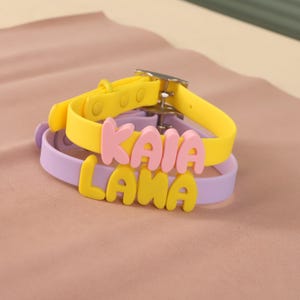 May include: Two silicone collars with metal buckles. The top yellow collar features pink raised letters spelling KAIA. The bottom lavender collar features yellow raised letters spelling LANA. Both collars have adjustable straps with circular holes.