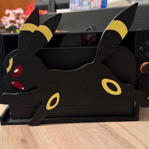 Puede incluir: Un soporte de consola de juegos Nintendo Switch de madera negra con un diseño Umbreon estilizado con detalles amarillos. El soporte tiene una base rectangular y una ranura para sujetar la consola. La figura de Umbreon tiene ojos rojos y una expresión amenazante.