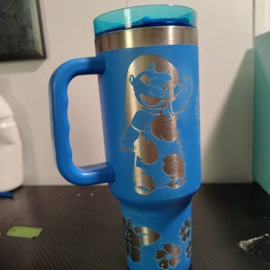 Peut inclure: Un gobelet bleu en acier inoxydable avec une poign&eacute;e et un couvercle bleu translucide. Le gobelet pr&eacute;sente un motif grav&eacute; au laser d'un personnage de dessin anim&eacute; aux grandes oreilles et un motif floral sur la partie inf&eacute;rieure en m&eacute;tal argent&eacute;.