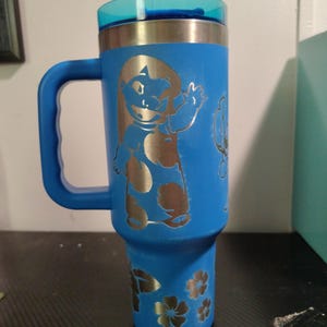 Peut inclure: Un gobelet isotherme bleu vif avec une poign&eacute;e et un couvercle bleu translucide. L'ext&eacute;rieur pr&eacute;sente un motif grav&eacute; au laser d'un personnage de dessin anim&eacute; en robe &agrave; motifs faisant signe, ainsi que des motifs floraux grav&eacute;s pr&egrave;s de la base.