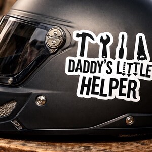 Könnte beinhalten: Schwarzer Motorradhelm mit klarem Visier. Ein weißer Aufkleber mit schwarzen Werkzeugen und dem Text "DADDY'S LITTLE HELPER" ist an der Seite angebracht. Der Helm liegt auf einer Holzoberfläche.