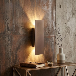 May include: A bronze-colored wall sconce with a rectangular design, emitting warm light. The sconce is mounted on a textured, rust-colored wall. A small table holds a leather-bound journal, a compass, and a vase with branches.