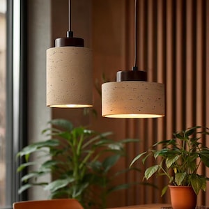 Peut inclure: Deux lampes suspendues cylindriques avec un abat-jour beige clair texturé et un dessus marron foncé. Les lumières sont suspendues à un cordon foncé, éclairant l'espace en dessous. La scène comprend une table en bois, des plantes et un mur en bois.