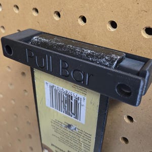 May include: A black plastic pull bar tool with the text Pull Bar embossed on the front. The tool features two circular mounting holes on the ends and is attached to a yellow product packaging card that displays a barcode and printed text.