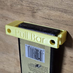 Pode incluir: Uma barra de puxar amarela com o texto "Pull Bar" impresso, segurando um rolo preto. A barra de puxar tem dois furos em cada lado. A barra de puxar está em cima de um objeto preto com um código de barras.