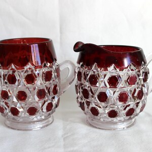 May include: A vintage sugar bowl and creamer set. The clear glass pieces have a ruby red top and ruby red hexagonal accents. The sugar bowl has two handles, and the creamer has a handle and a spout.