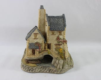 May include: A miniature stone cottage figurine with a gray shingled roof, a tall chimney, and a small arched bridge. The cottage is adorned with small windows, doors, and colorful floral accents. The base is textured to resemble a rocky landscape.