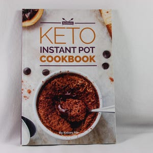 May include: A cookbook titled "Keto Instant Pot Cookbook" by Kelsey Ale. The cover features a close-up of a brown chocolate dessert in a white bowl with a spoon.