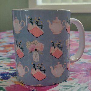 Può includere: Tazza in ceramica con sfondo azzurro chiaro con un motivo ripetuto di teiere bianche, libri rosa e mazzi di fiori. La tazza ha un manico bianco ed è appoggiata su una tovaglia floreale.