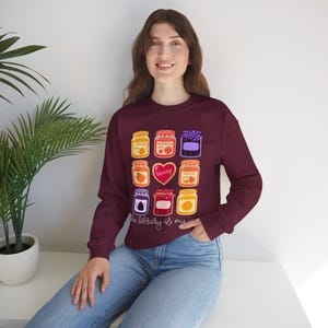 Pode incluir: Camisola bordô com ilustrações coloridas de potes de geleia. Os potes são rotulados com nomes de frutas e o texto "the library is my jam". O design inclui potes de geleia de laranja, morango e pêssego.