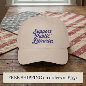 May include: A beige baseball cap featuring the embroidered blue text Support Public Libraries with small red and blue stars. The cap has a curved brim and a structured crown, displaying a clean design centered on the front panel of the hat.
