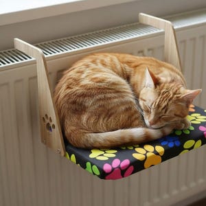 Könnte beinhalten: Eine schlafende, orangefarbene getigerte Katze auf einem Katzenbett, das an einem Heizkörper befestigt ist. Das Bett hat ein schwarzes Kissen mit bunten Pfotenabdrücken. Der Holzrahmen hat Pfotenabdruck-Ausschnitte.