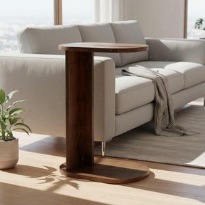 Puede incluir: Una mesa auxiliar en forma de C de madera marrón oscuro con una superficie y base ovaladas. La mesa tiene un soporte vertical y está colocada junto a un sofá gris claro. También se ven una planta en maceta y una alfombra.