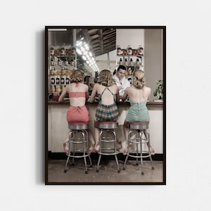 May include: A framed vintage print depicts three women in retro swimsuits seated at a diner counter. The women are seated on chrome stools, facing a bartender. The bar is stocked with bottles and drinks. The print has a sepia tone.