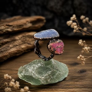 May include: A ring featuring a dark, textured metal band with an open design, topped with two raw, uncut gemstones: one flat, light blue stone and one smaller, vibrant pink stone with dark inclusions, both set in a dark, metallic frame.