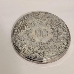May include: A round silver-toned metal compact case featuring an intricate embossed floral and scrollwork pattern across the surface. The center of the lid displays a circular smooth area engraved with the stylized monogram letters WHB in cursive script.