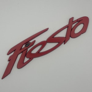 Pode incluir: Um emblema vermelho "Fiesta", provavelmente para um veículo, é exibido em um fundo branco. A palavra está em uma fonte estilizada, com um acabamento brilhante. O emblema está ligeiramente inclinado para a esquerda.