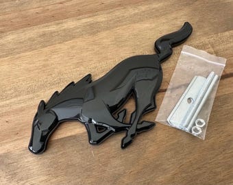 May include: A glossy black automotive emblem shaped like a galloping horse in a running pose. The emblem is accompanied by a small clear plastic bag containing two silver-colored metal mounting brackets and two matching hexagonal nuts.
