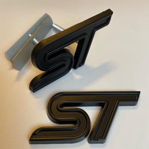 May include: Black ST emblem featuring a stylized S and T design. The emblem is made of a matte black material. A silver metal bracket with a screw is visible in the background.