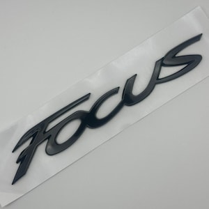 May include: Black "FOCUS" emblem on a white backing. The emblem is in a stylized font, with a glossy finish. The emblem is likely for a vehicle.