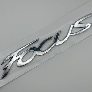 Puede incluir: Un emblema cromado "Focus", probablemente para un vehículo, se muestra sobre un respaldo blanco. La escritura del emblema está en una fuente estilizada, reflejando la luz y pareciendo tridimensional. El fondo es una superficie gris claro lisa.