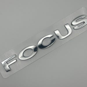 Puede incluir: Una taza de cerámica blanca con un asa negra y el texto "But First, Coffee" en negro, con una fuente de estilo manuscrito. La taza está sobre una superficie blanca.