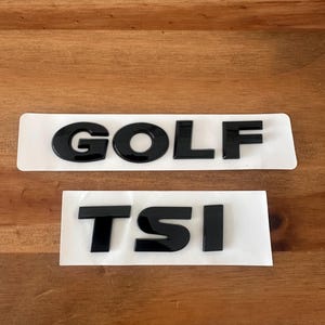 Puede incluir: Dos emblemas de coche negros sobre un respaldo blanco. El emblema superior deletrea "GOLF" en letras negritas y brillantes. Debajo, las letras "TSI" se muestran en un estilo similar. Los emblemas están sobre un fondo de madera.