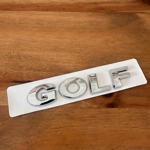 May include: Chrome "GOLF" emblem on a white backing. The letters are shiny and reflective, with a rounded font. The emblem is placed on a wooden surface.