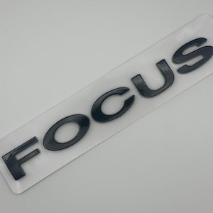 Może przedstawiać: Czarny emblemat "FOCUS" na białym prostokątnym podkładzie. Litery są pogrubione, bezszeryfowe, z błyszczącym wykończeniem. Emblemat jest prawdopodobnie przeznaczony do użytku motoryzacyjnego.