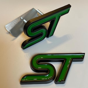 May include: Two green and black ST emblems with a metallic mounting bracket. The emblems have a glossy finish and a raised design. The letters are outlined in black and filled with a vibrant green colour. The background is a plain white surface.