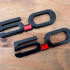 Puede incluir: Dos emblemas negros "5.0" con un acento rojo, probablemente para un vehículo. Los emblemas están hechos de un material brillante y se muestran sobre una superficie de madera. El "5" está estilizado con una fuente distintiva.