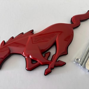 May include: A red, glossy Mustang horse emblem with black trim, on a white surface. The emblem is in a running pose. Includes mounting hardware in a small plastic bag.
