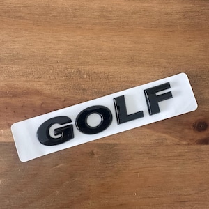 Puede incluir: Emblema "GOLF" negro brillante sobre un respaldo rectangular blanco. Las letras son en negrita y destacan sobre el fondo blanco. El emblema es probablemente para un vehículo y está sobre una superficie de madera.