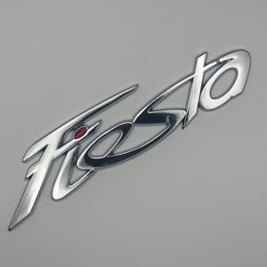 Pode incluir: Um emblema cromado "Fiesta" com um detalhe vermelho. A palavra está em uma fonte de escrita estilizada, com um design moderno. O fundo é uma superfície cinza clara lisa.