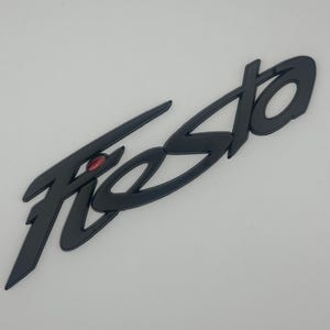 Pode incluir: Emblema preto "Fiesta" com um pequeno detalhe vermelho. O emblema é uma fonte de script estilizada, provavelmente para um veículo. O emblema é feito de material preto fosco sobre um fundo branco.
